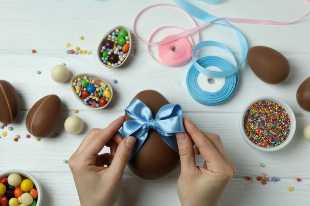 Weibliche Hände halten Bogen auf Schokoladenei auf weißer Holzoberfläche mit Osterzubehör