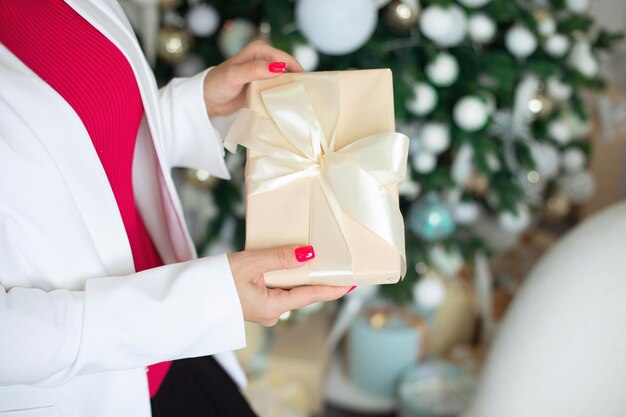 Foto weibliche hände halten eine geschenkkiste, die mit einem goldenen band auf dem hintergrund eines weihnachtsbaums geschmückt ist