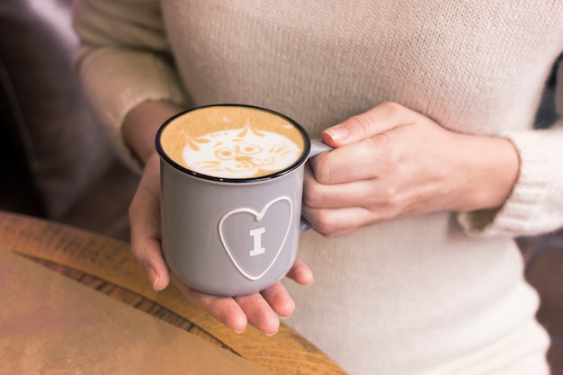 Weibliche Hände halten eine graue warme emaillierte Tasse mit einem Herzsymbol.