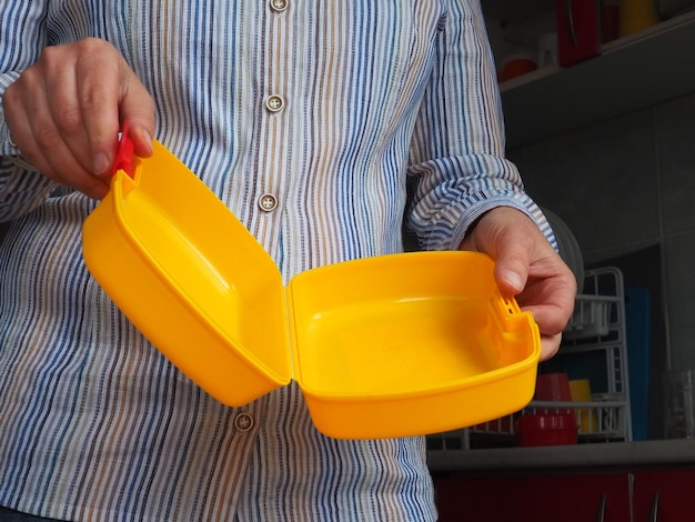 Weibliche Hände halten einen gelben, offenen Plastikbehälter oder eine Verpackung für das Mittagessen in der Schule Küchenutensilien Eine Frau in einem weiß gestreiften Hemd zeigt das Thema Lunchbox Lebensmittelhygiene und Recycling