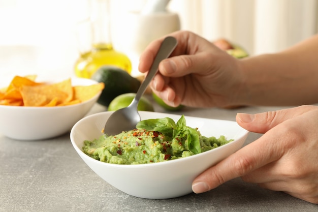 Weibliche Hände halten Löffel und Schüssel mit Guacamole auf grauem Tisch