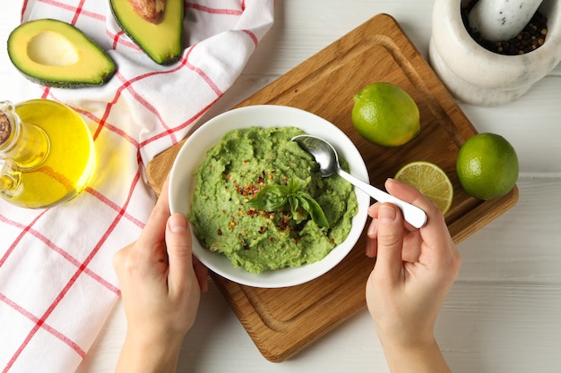 Weibliche Hände halten Löffel und Schüssel mit Guacamole auf weißem Holztisch