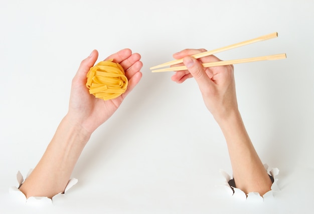 Weibliche Hände halten rohe Tagliatelle-Nudeln und Essstäbchen auf Weiß mit zerrissenem Loch. Minimalistisches Lebensmittelkonzept. Draufsicht