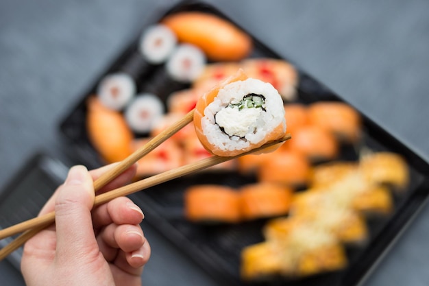 Weibliche Hände halten Sushi-Rollen mit Stöcken nahe, verschwommener dunkler Hintergrund