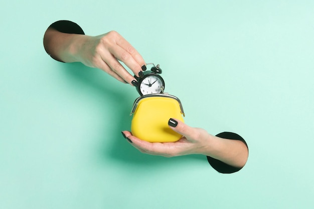 Weibliche Hände halten Wecker und Brieftasche durch ein Loch auf Neon-Minz-Hintergrund Zeit ist Geld-Konzept
