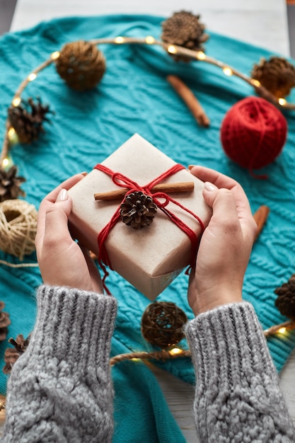 Weibliche Hände halten Weihnachtsgeschenkbox gegen rote Socken, einen blauen Strickpullover und eine Girlande mit Zapfen. Übergibt ein Geschenk
