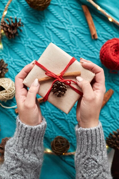 Weibliche Hände halten Weihnachtsgeschenkbox gegen rote Socken, einen blauen Strickpullover und eine Girlande mit Zapfen