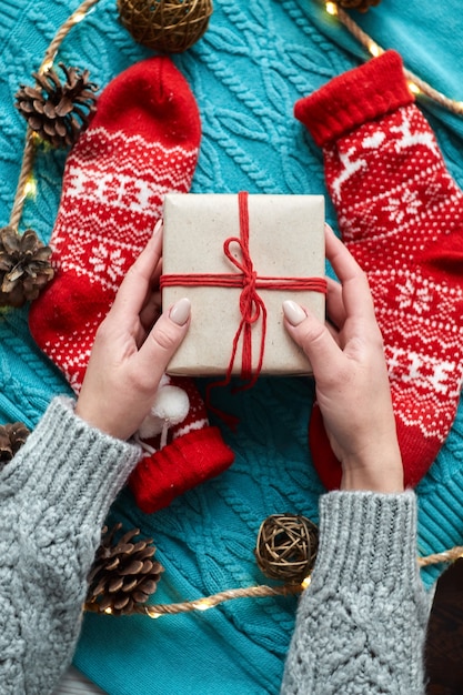 Weibliche Hände halten Weihnachtsgeschenkbox und rote Socken, einen blauen Strickpullover und eine Girlande mit Zapfen. Draufsicht