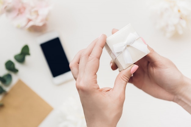 Weibliche Hände halten weiße Geschenkbox auf Blumen und Telefon. Draufsicht