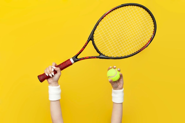 Weibliche Hände heben und halten einen Tennisschläger und einen Tennisball auf einem gelben isolierten Hintergrund