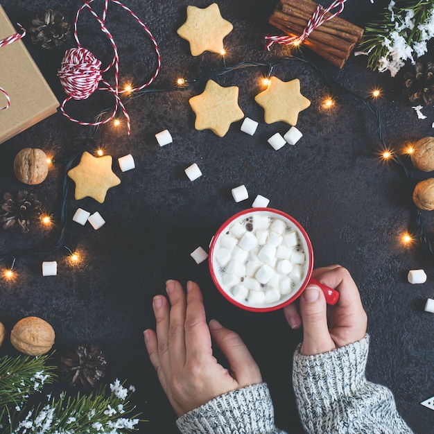 Weibliche Hände in der Strickjacke halten Becher Kakao mit Eibisch- und Weihnachtsdekorationen