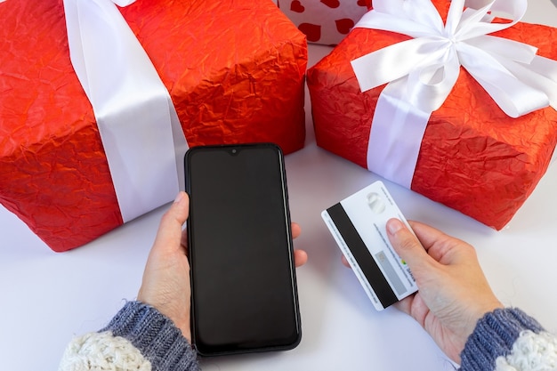 Weibliche Hände in einem Pullover halten ein Smartphone und eine Zahlungskarte in der Nähe der Geschenkboxen auf dem Tisch. Konzept des festlichen Online-Shoppings für Weihnachten und Neujahr