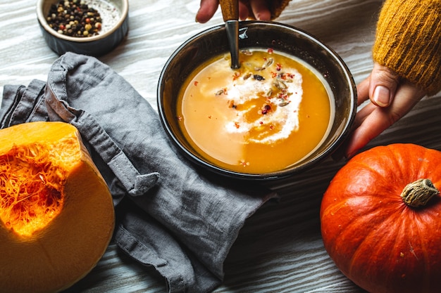 Weibliche Hände in gelbem Strickpullover, die eine Schüssel mit Kürbiscremesuppe auf dunklem Steinhintergrund mit Löffel mit geschnittenem frischem Kürbis, Draufsicht halten. Herbstliches gemütliches Abendessen Konzept