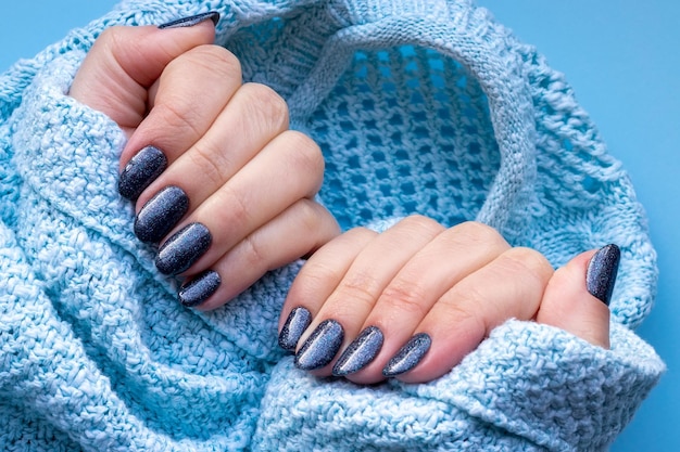 Weibliche Hände in gestricktem Pulloverstoff mit schöner Maniküre dunkelblau glitzernden Nägeln auf blauem Hintergrund Selektiver Fokus Detailansicht