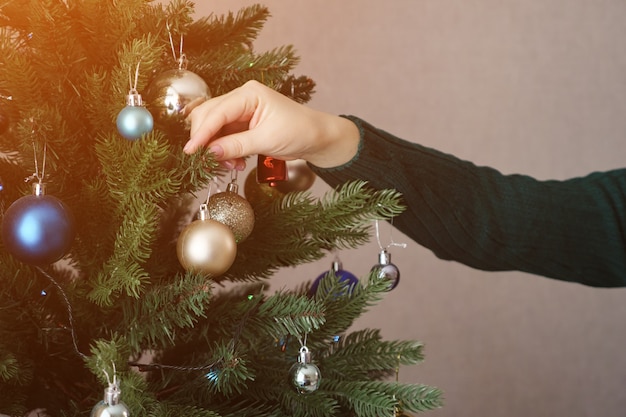Weibliche Hände in grünem Pullover halten und hängen goldenen Spielzeugball am künstlichen Weihnachtsbaum mit bunten und funkelnden Spielzeugbällen