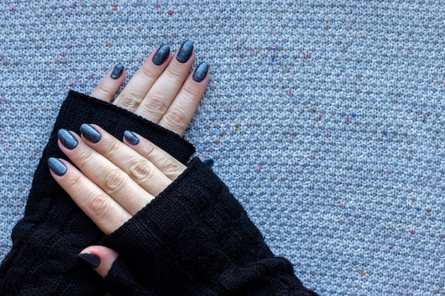 Weibliche Hände in schwarzen Strickhandschuhen mit schöner Maniküre - dunkelblaue glitzernde Nägel auf gestricktem Hintergrund mit Kopierraum