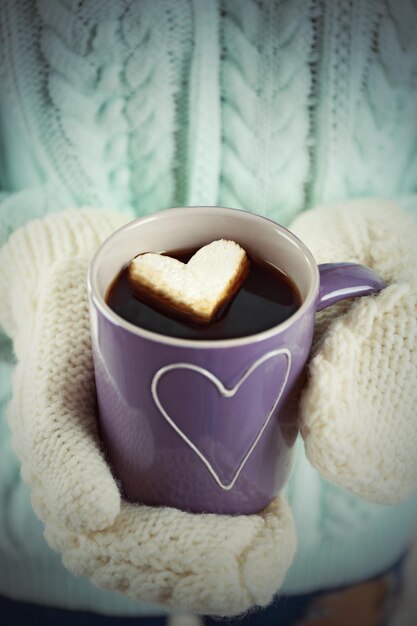 Foto weibliche hände in warmen fäustlingen, die eine tasse heißen kaffee mit herz-marshmallow in der nähe halten