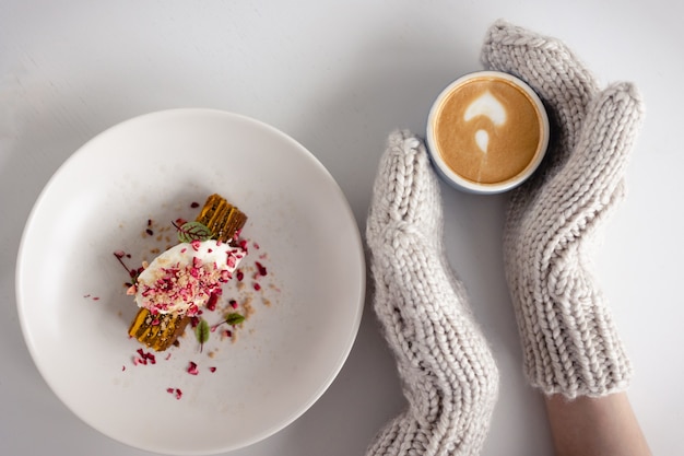 Weibliche Hände in weißen Handschuhen halten Tasse heißen Kaffees mit Schaum auf weißem Tisch mit Kuchen. Weihnachtshintergrund. Konzept von Winter, Wärme, Feiertagen, Ereignissen. Weicher Fokus. Draufsicht.