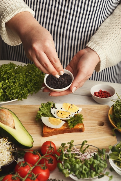 Weibliche Hände machen Bruschettas mit Eiern, Salat und schwarzem Sesam. Gesunde vegetarische Sandwiches am Küchentisch mit verschiedenen Gemüseschüsseln und Grüns. Kochen Essen Hintergrund, Nahaufnahme