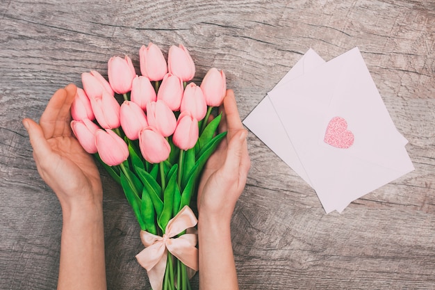 Weibliche hände mit einem blumenstrauß von rosa tulpen und leeren weißen umschlägen für buchstaben, auf einem hölzernen hintergrund.