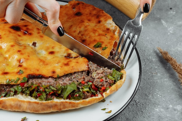 Weibliche Hände mit einem Messer und einer Gabel schneiden Khachapuri mit Lamm und Chili