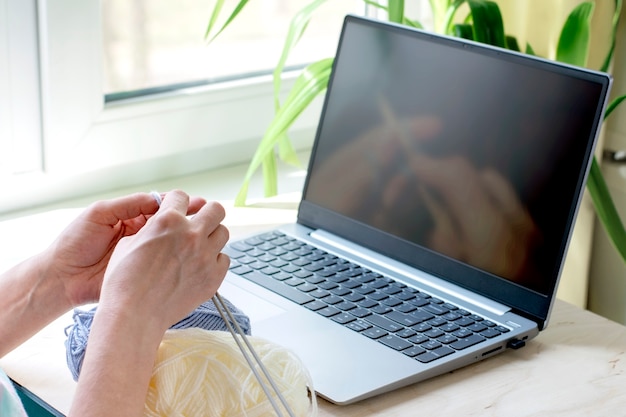 Weibliche Hände mit Fäden und Stricknadeln. Ein Mädchen sieht sich Lehrvideos zum Stricken auf einem Laptop an.