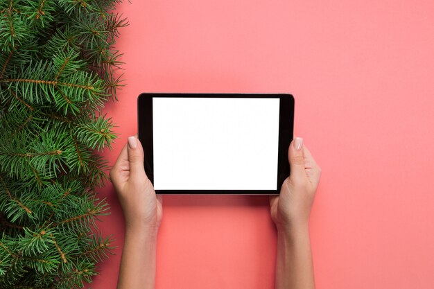 Weibliche Hände mit Tablet-Computer auf rosa Weihnachtshintergrund. Draufsicht des Weihnachtsbaumasts