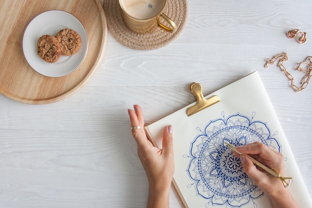 Weibliche Hände schließen oben zeichnen dekoratives rundes Blumenmandala. Hobby und Entspannung zu Hause. Eine Tasse Kaffee und Kekse auf einem Holztablett. Weißer Hintergrund.