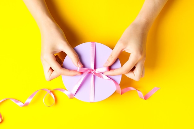 Weibliche Hände und runde Geschenkbox mit Schleife auf gelbem Hintergrund