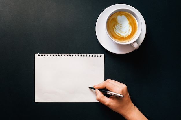 Weibliche hand, die auf weißem papier mit einer tasse latte-kaffee auf dem holzschreibtisch aufschreibt, ansicht von oben