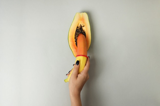 Weibliche Hand, die Banane mit Kondom in Papaya auf hellgrauem Hintergrund hält