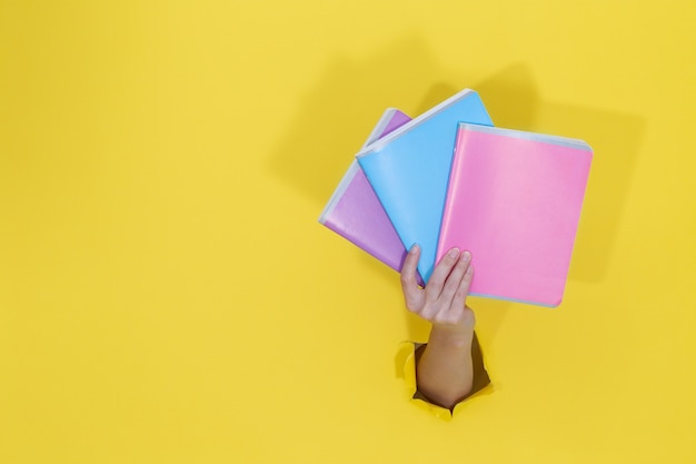 Weibliche Hand, die bunte Notizbücher vom gelben zerrissenen Papier hält. Back to School Bildungskonzept. Schulsachen und Büromaterial. Platz für Text.