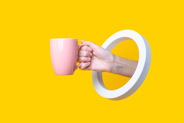 Weibliche Hand, die eine rosa Tasse in einem runden Loch auf einem gelben Hintergrund hält.