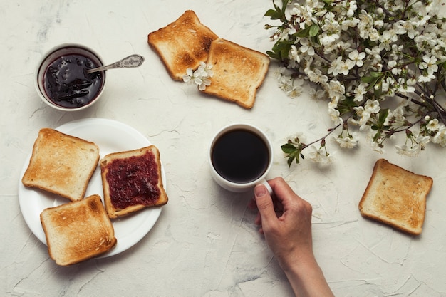 Weibliche Hand, die eine Tasse Kaffee, Marmelade, Toast, Frühlingszweigbaum mit Blumen hält. Frühstückskonzept. Flache Lage, Draufsicht