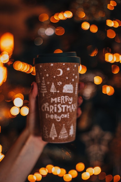 Weibliche Hand, die eine Tasse Kaffee oder Tee mit einem Neujahrs- und Weihnachtsdesign hält Winterurlaubsdekor