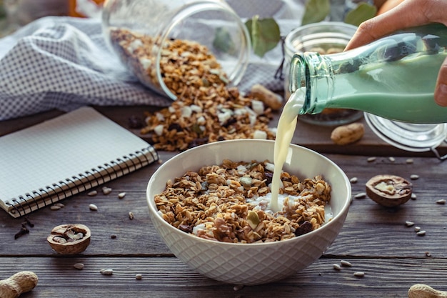 Weibliche Hand, die Glasflasche hält, die Milch in Müsli-Müsliflocken-Schüssel gießt