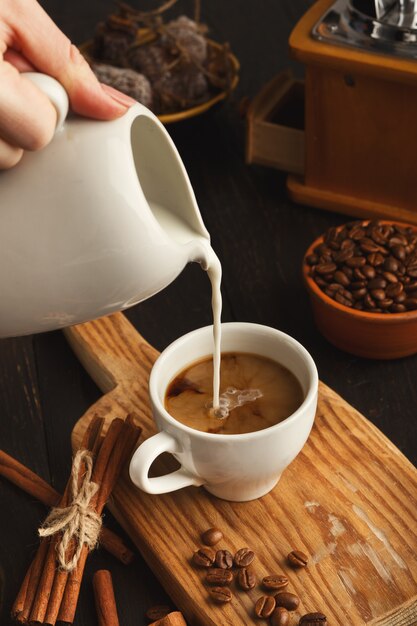 Weibliche Hand, die Morgenkaffee zum Frühstück macht. Milch in Espresso gießen, Nahaufnahme. , Weiße Tasse und Zimt auf altem rustikalem Holzbrett, Kopierraum