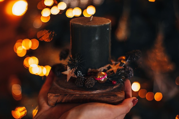 Weibliche Hand, die Weihnachtskomposition mit Kerze vor einem Hintergrund von festlichen Lichtern hält