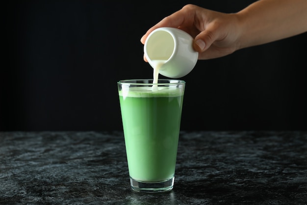 Weibliche Hand gießt Milch in ein Glas Matcha Latte vor schwarzem Hintergrund