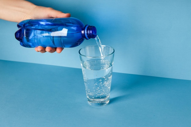 Weibliche Hand gießt Wasser in Glas auf blauem Hintergrund