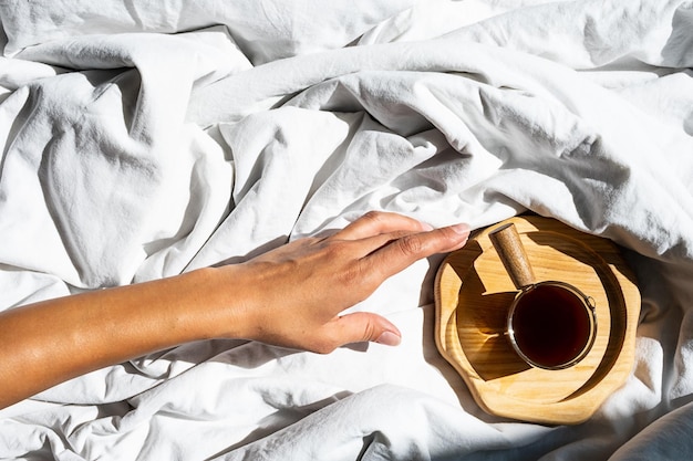 Weibliche Hand greift nach morgendlicher Kaffeetasse im Bett Frau wacht auf weißer Bettwäsche im Sonnenlicht auf