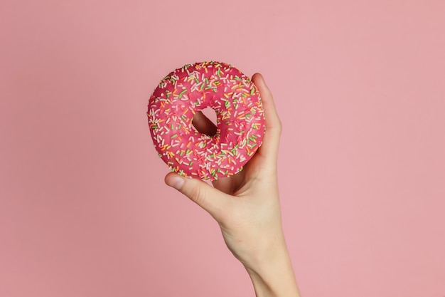Weibliche Hand hält einen glasierten Donut auf rosa Hintergrund. Pastellfarbentrend. Gewichtsverlust nach den Ferien. Draufsicht.