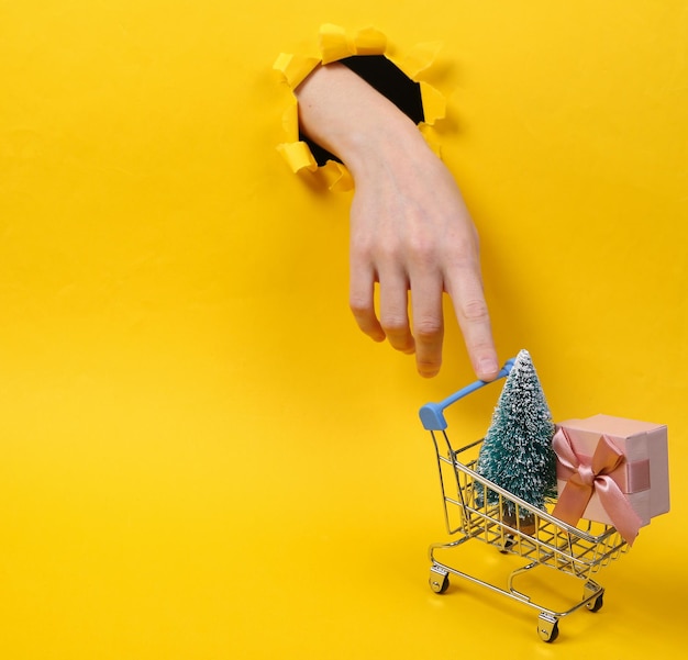 Weibliche Hand hält Einkaufswagen mit Weihnachtsbaum und Geschenkbox durch zerrissenes Loch gelbes Papier Konzeptkunst Minimalismus