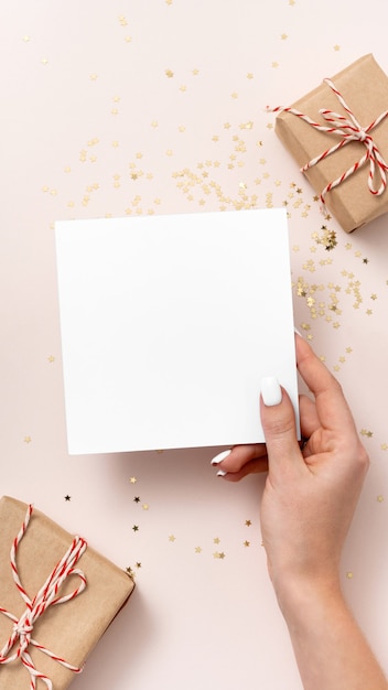 Weibliche Hand hält leeres quadratisches Papiermodell, Konfetti mit goldenen Sternen, Geschenkboxen auf beigem Hintergrund. Flache Lage, Draufsicht, Kopierraum, minimalistisch. Weihnachts- und Neujahrskonzept