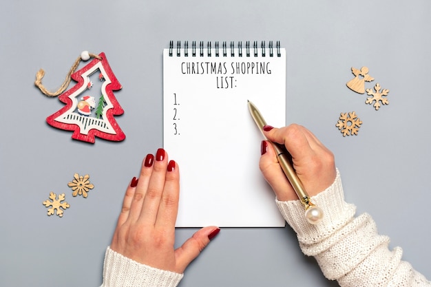 Weibliche Hand hält Stift, Weihnachtseinkaufsliste schreibend, Geschenkideen auf weißem Notizblock auf grau