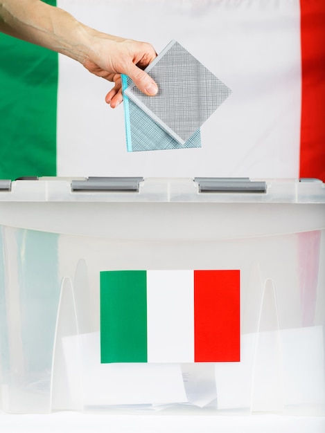 Weibliche Hand hält Stimmzettel über Wahlurne. Italienische Flagge im Hintergrund. Nahaufnahme