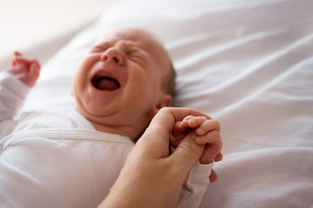 Weibliche Hand hält weinendes neugeborenes Baby, das auf dem Bett liegt, Konzept der Liebe, Pflege, Schutz, Mutterunterstützung