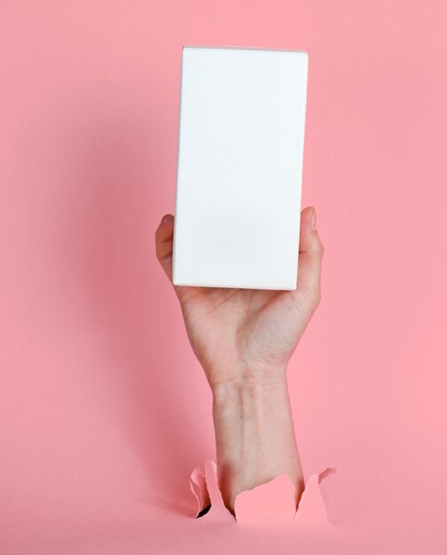 Weibliche Hand hält weiße Box durch zerrissenes rosa Papier. Minimalistisches kreatives Modekonzept