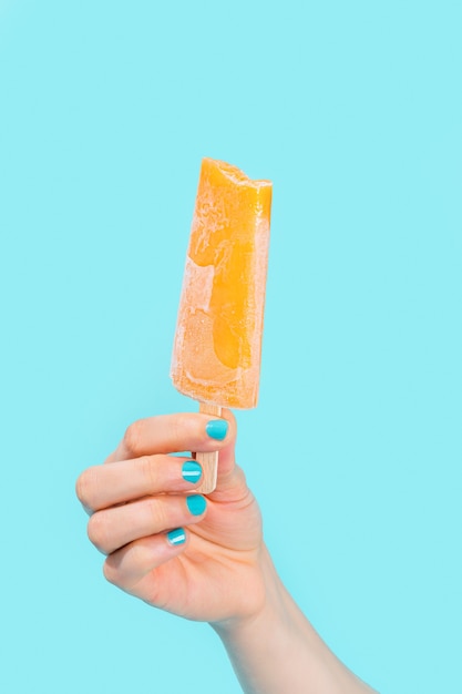 Weibliche Hand mit blauem Nagellack, die orange Eiscreme auf blauem Hintergrund hält