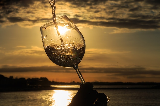 Weibliche Hand mit einem Glas Weißwein mit schönem Sonnenuntergang
